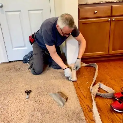 Chris kneeling at a threshold, actively stretching carpet with a knee kicker and utility knife on the floor beside him
