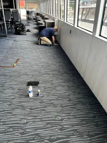 A technician kneeling at a window wall aboard a commercial tour boat, patterned carpet underfoot